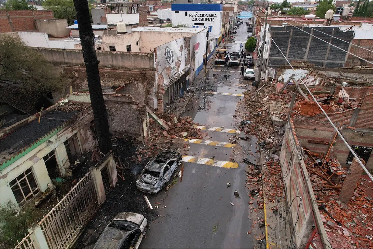 Toma aérea de una calle con severos daños estructurales, escombros de ladrillo sobre el asfalto, fachadas derrumbadas y dos automóviles calcinados tras una explosión en Ojocaliente.