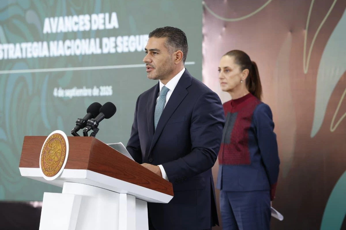 Omar García Harfuch hablando desde un pódium oficial de madera con el sello nacional mientras Claudia Sheinbaum observa de pie al fondo en una presentación sobre avances de seguridad.
