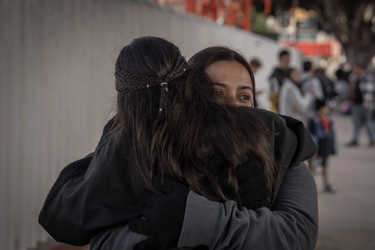 Primer plano de dos mujeres dándose un abrazo cálido al aire libre, con una de ellas sonriendo levemente sobre el hombro de la otra mientras la rodea con sus brazos vestidos con guantes negros.