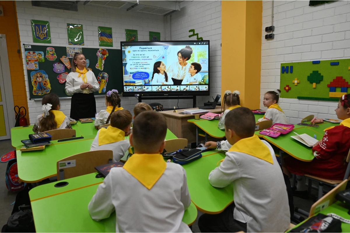 Profesora frente a un grupo de estudiantes sentados en pupitres verdes, mostrando una lección en una pantalla digital interactiva dentro de un salón de clases equipado con tecnología.