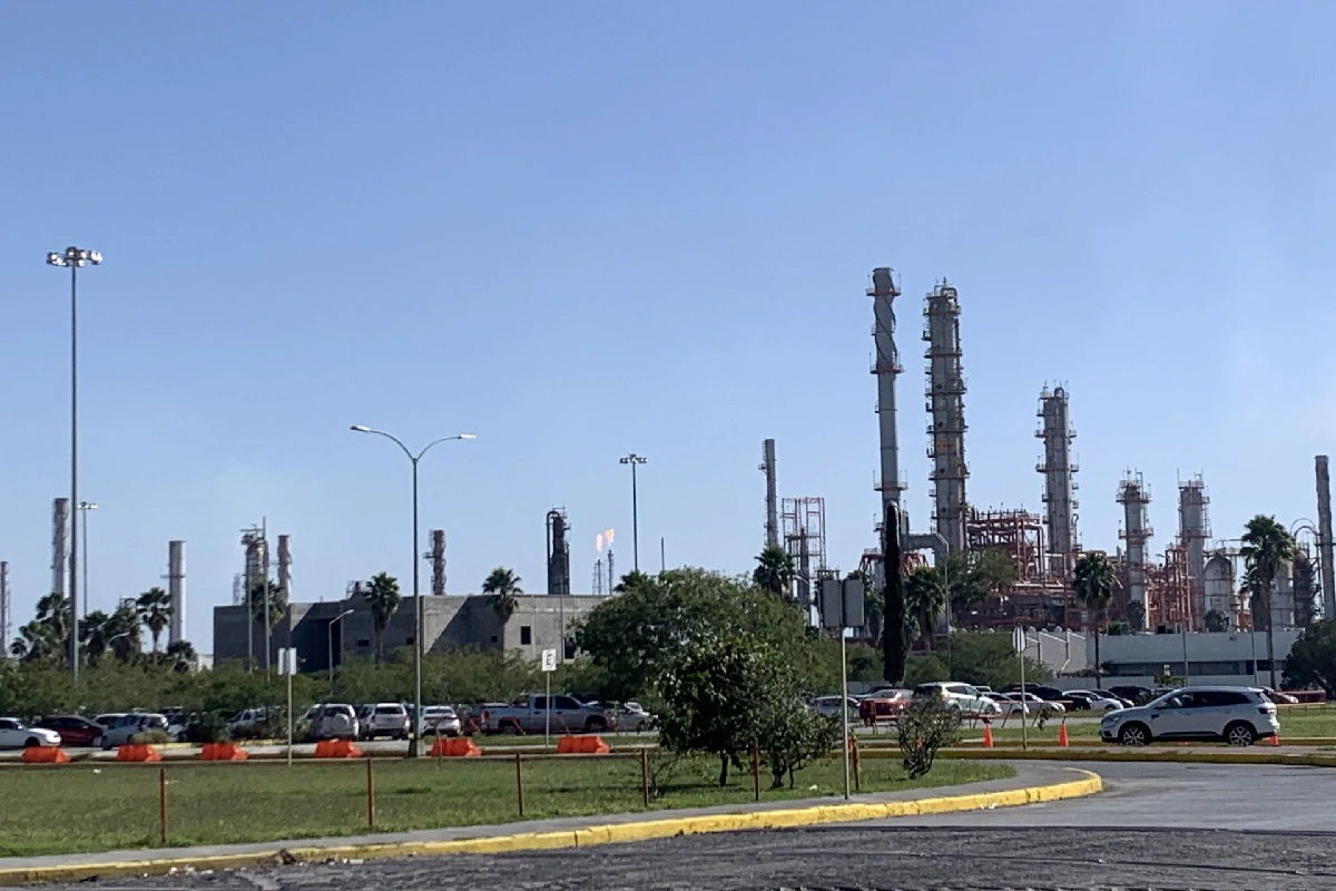 Vista exterior de las instalaciones industriales de la refinería de Cadereyta con torres de procesamiento, chimeneas con llamas encendidas y estacionamiento bajo un cielo despejado.
